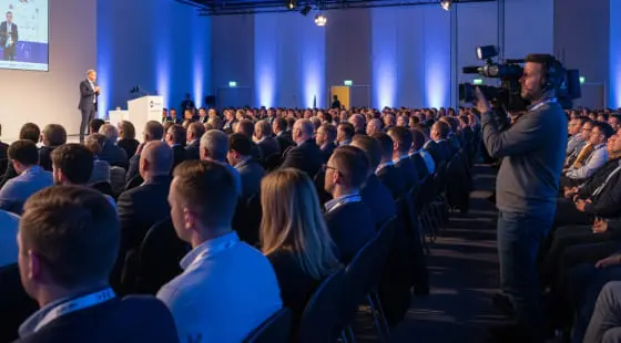 Оператор снимает зал с участниками конференции в деловой одежде и спикером на сцене, кадр подготовлен для Видеосъемки конференции