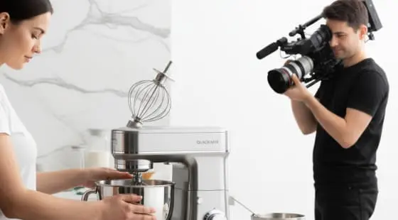 Женщина в белой футболке использует настольный миксер, оператор с камерой снимает ее крупным планом на светлом фоне с мраморной стеной, видеосъемка для интернет-магазина